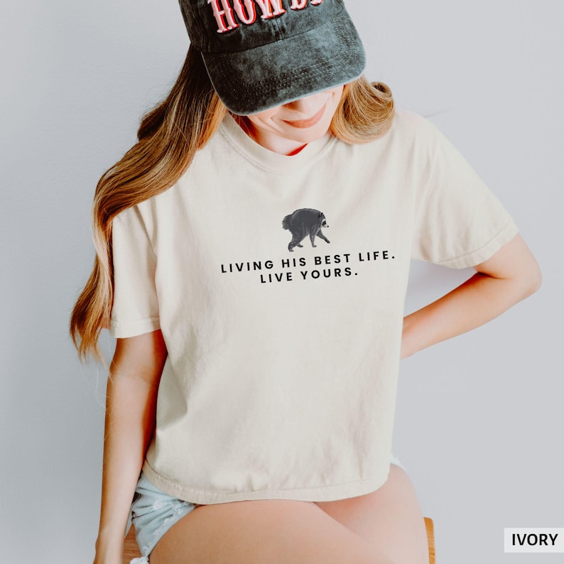 May include: An ivory t-shirt featuring a small graphic of a raccoon and the black text "LIVING HIS BEST LIFE. LIVE YOURS." The shirt has short sleeves and a crew neckline. A dark grey baseball cap with red lettering is worn above the shirt.