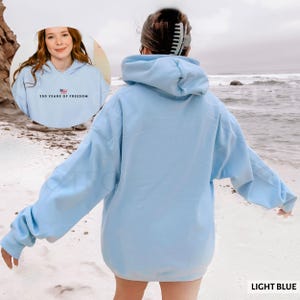 Puede incluir: Sudadera con capucha azul claro con el texto "250 YEARS OF FREEDOM" y un peque&ntilde;o gr&aacute;fico de la bandera estadounidense. La sudadera tiene una capucha con cord&oacute;n y mangas largas. Las palabras "LIGHT BLUE" est&aacute;n impresas en la esquina inferior derecha.