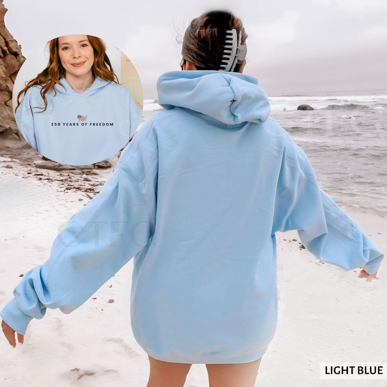 Puede incluir: Sudadera con capucha azul claro con el texto "250 YEARS OF FREEDOM" y un peque&ntilde;o gr&aacute;fico de la bandera estadounidense. La sudadera tiene una capucha con cord&oacute;n y mangas largas. Las palabras "LIGHT BLUE" est&aacute;n impresas en la esquina inferior derecha.