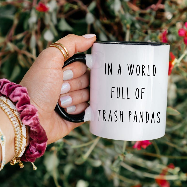 May include: A white ceramic mug with a black handle and interior, featuring the black printed text IN A WORLD FULL OF TRASH PANDAS. The mug has a smooth, glossy finish and a cylindrical shape, designed for holding hot or cold beverages.
