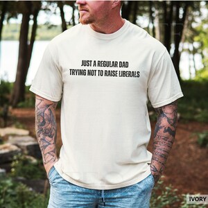 May include: Ivory-colored t-shirt with the text "JUST A REGULAR DAD TRYING NOT TO RAISE LIBERALS" in black. The shirt is short-sleeved and worn by a person with tattoos on their arms. The background is a blurred outdoor scene.