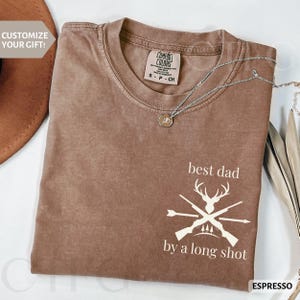 May include: Espresso-colored t-shirt with the text "best dad by a long shot" and a graphic of crossed rifles, arrows, and deer antlers. A silver necklace with a pendant is draped across the shirt. The shirt has a Comfort Colors tag.