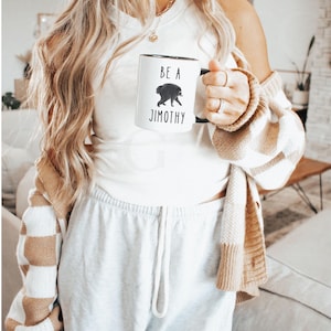 May include: A white ceramic mug with a black handle featuring a graphic illustration of a raccoon and the black text "BE A JIMOTHY" printed on the front. The mug has a cylindrical shape and a smooth finish, displaying the text and animal clearly.