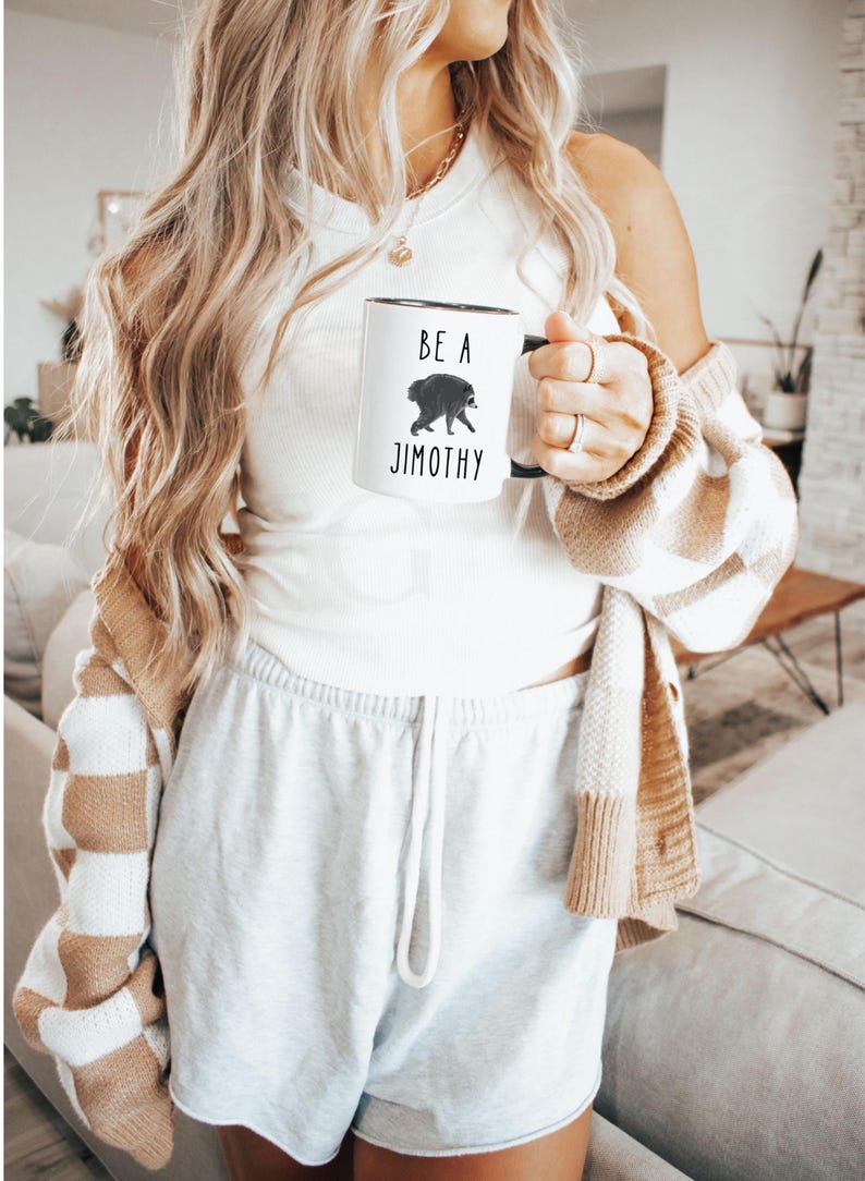 May include: A white ceramic mug with a black handle featuring a graphic illustration of a raccoon and the black text "BE A JIMOTHY" printed on the front. The mug has a cylindrical shape and a smooth finish, displaying the text and animal clearly.