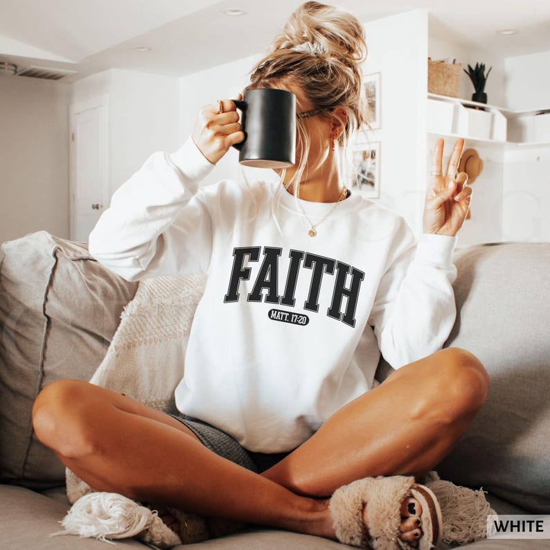 Puede incluir: Sudadera blanca con la palabra "FAITH" en letras negras, con "MATT. 17:20" debajo. La sudadera es de manga larga y tiene cuello redondo. La persona sostiene una taza negra.