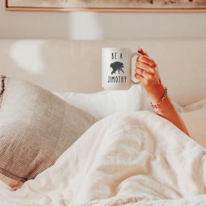 May include: A white ceramic mug featuring a black graphic of a raccoon and the text "BE A JIMOTHY" printed in a simple, dark font. The mug has a curved handle on the right side and a smooth, cylindrical body, designed for holding hot or cold beverages.