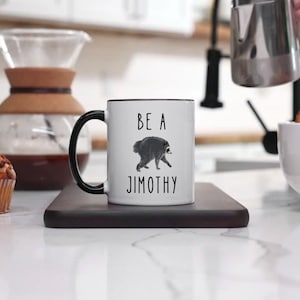 May include: A white ceramic mug with a black handle and rim, featuring a black graphic of a raccoon and the text BE A JIMOTHY printed in black. The mug is centered on a dark rectangular coaster, showcasing a clean and simple design.