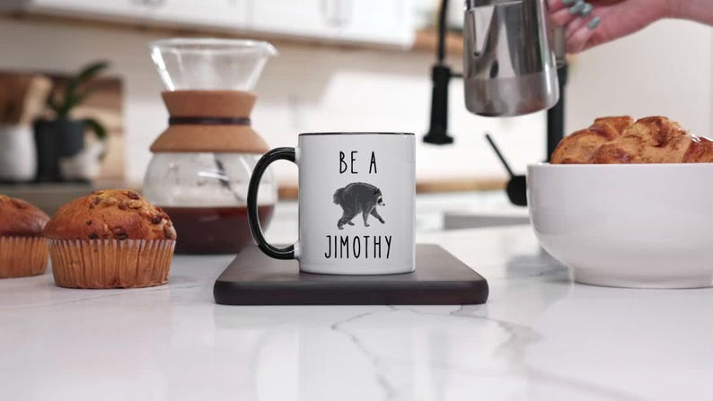 May include: A white ceramic mug with a black handle and rim, featuring a black graphic of a raccoon and the text BE A JIMOTHY printed in black. The mug is centered on a dark rectangular coaster, showcasing a clean and simple design.