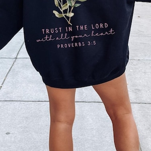 May include: Black hoodie with a floral design and the text "TRUST IN THE LORD with all your heart PROVERBS 3:5". The image also includes the text "BEST CHRISTIAN APPAREL". The hoodie is paired with white high-top sneakers.