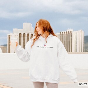 Puede incluir: Una sudadera con capucha blanca con el texto "250 YEARS OF FREEDOM" y una peque&ntilde;a bandera estadounidense. La sudadera tiene un bolsillo delantero y la lleva una persona.