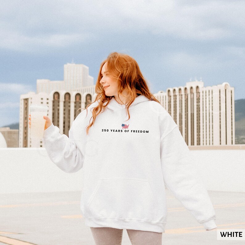 Puede incluir: Una sudadera con capucha blanca con el texto "250 YEARS OF FREEDOM" y una peque&ntilde;a bandera estadounidense. La sudadera tiene un bolsillo delantero y la lleva una persona.