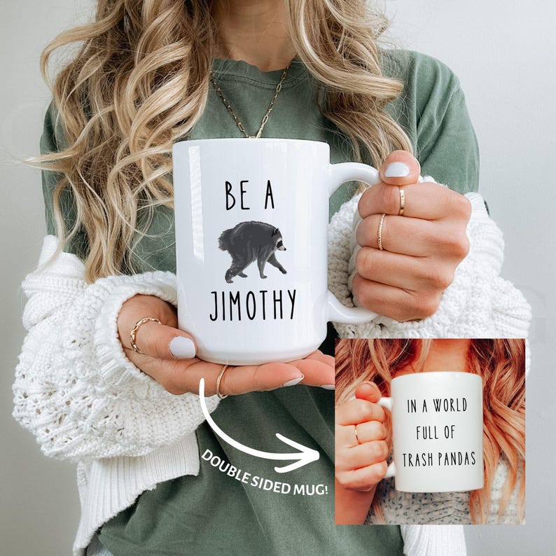May include: A white ceramic mug featuring a black graphic of a raccoon and the text BE A JIMOTHY on one side. The reverse side displays the text IN A WORLD FULL OF TRASH PANDAS. The mug has a curved handle and is shown in a double-sided view.