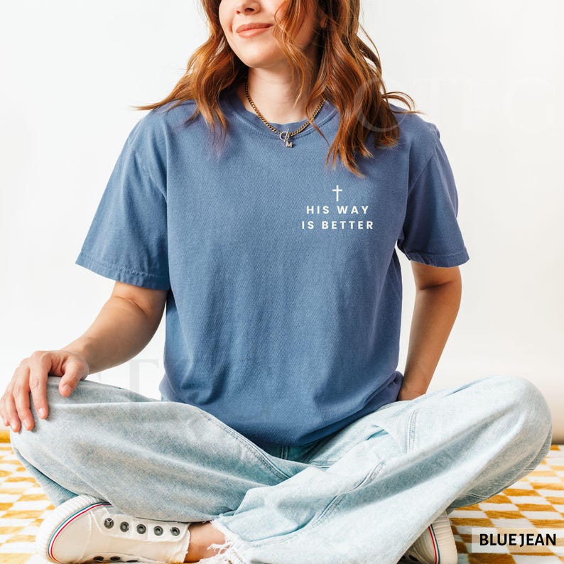 May include: A blue t-shirt with the text "HIS WAY IS BETTER" and a cross graphic. The shirt is a solid color and has short sleeves. The person is wearing light blue jeans.