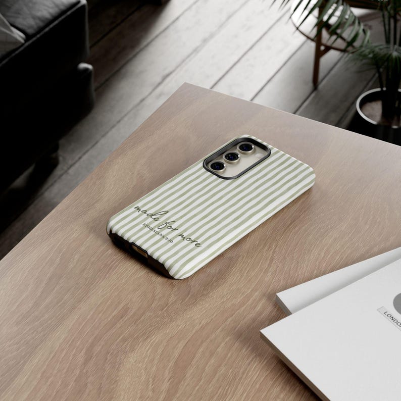 May include: A smartphone case with a striped pattern in shades of green and white. The case has a black camera module and the words "made for more" printed on it. The case is lying on a wooden surface.