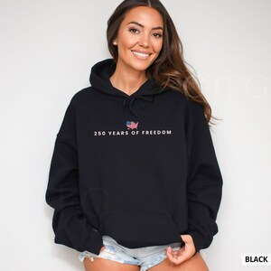 Puede incluir: Sudadera con capucha negra con el texto "250 YEARS OF FREEDOM" y un gr&aacute;fico de la bandera estadounidense dentro del contorno de los Estados Unidos. La sudadera tiene una capucha con cord&oacute;n y un bolsillo delantero.