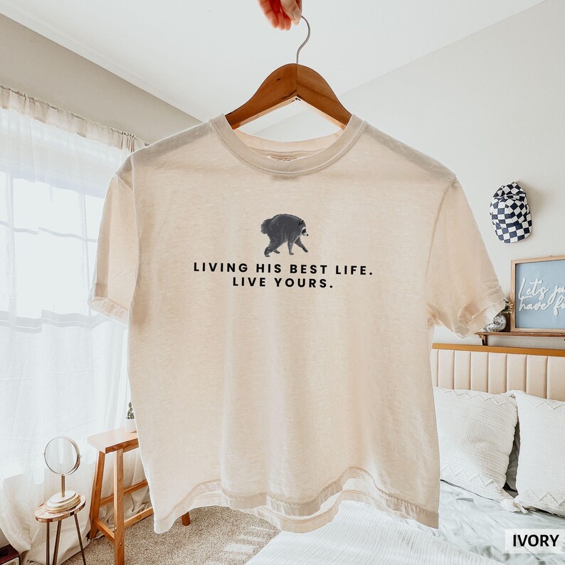 May include: An ivory-colored short-sleeve t-shirt featuring a graphic of a walking raccoon centered above the black text that reads, "LIVING HIS BEST LIFE. LIVE YOURS." The shirt is displayed on a wooden hanger against a bright, neutral indoor setting.