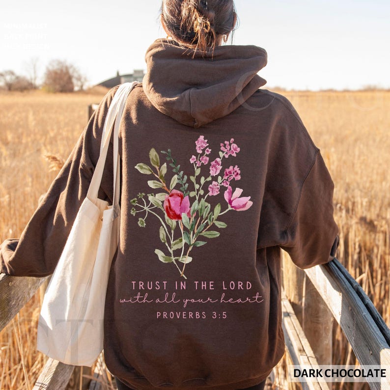 May include: Dark chocolate-colored hoodie with a floral design on the back, featuring pink and red flowers with green leaves. The text "TRUST IN THE LORD with all your heart PROVERBS 3:5" is printed below the flowers. A white tote bag is visible.