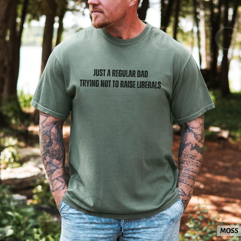 May include: Moss green t-shirt with the text "JUST A REGULAR DAD TRYING NOT TO RAISE LIBERALS" in black. The shirt is a crew neck style and appears to be made of a soft, comfortable material. The model is wearing blue jeans.