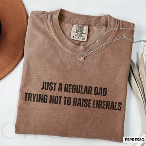 May include: Espresso-colored t-shirt with the text "JUST A REGULAR DAD TRYING NOT TO RAISE LIBERALS" in black. The shirt has a crew neck and short sleeves. A silver necklace with a small pendant is draped across the shirt. A brown hat is partially visible.