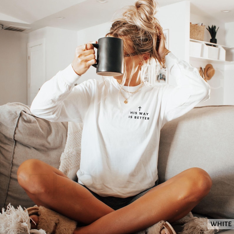 May include: A white long-sleeve shirt with the text "HIS WAY IS BETTER" and a cross graphic. The shirt is made of a soft material and is suitable for casual wear. A black mug is held up to the face.