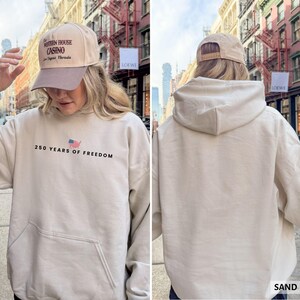 Puede incluir: Una sudadera con capucha color arena con un bolsillo delantero y el texto "250 YEARS OF FREEDOM" con una bandera estadounidense. La persona lleva una gorra de b&eacute;isbol beige con "The Western House Casino Las Vegas, Nevada" impreso.