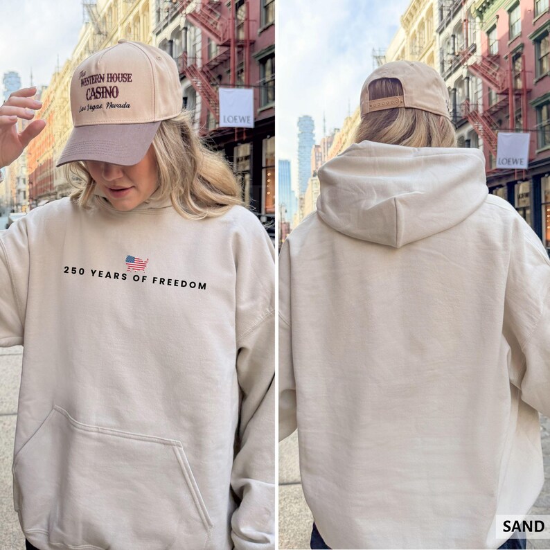 Puede incluir: Una sudadera con capucha color arena con un bolsillo delantero y el texto "250 YEARS OF FREEDOM" con una bandera estadounidense. La persona lleva una gorra de b&eacute;isbol beige con "The Western House Casino Las Vegas, Nevada" impreso.