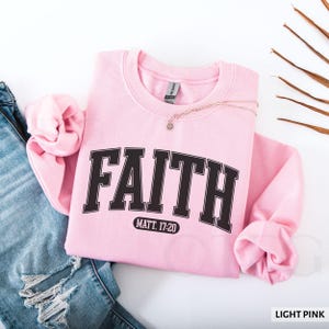 Puede incluir: Una sudadera rosa claro con la palabra "FAITH" en letras negras en negrita. Debajo est&aacute; el texto "MATT. 17:20". Un collar dorado con un colgante est&aacute; sobre la sudadera. Las palabras "LIGHT PINK" est&aacute;n en la esquina inferior derecha.