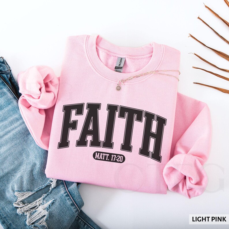 Puede incluir: Una sudadera rosa claro con la palabra "FAITH" en letras negras en negrita. Debajo est&aacute; el texto "MATT. 17:20". Un collar dorado con un colgante est&aacute; sobre la sudadera. Las palabras "LIGHT PINK" est&aacute;n en la esquina inferior derecha.