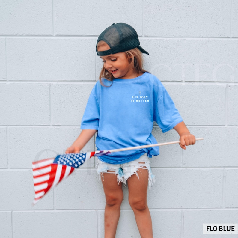 May include: A light blue t-shirt with the text "HIS WAY IS BETTER" and a cross graphic. The shirt is paired with denim shorts and a black mesh trucker hat. The person is holding a small American flag.