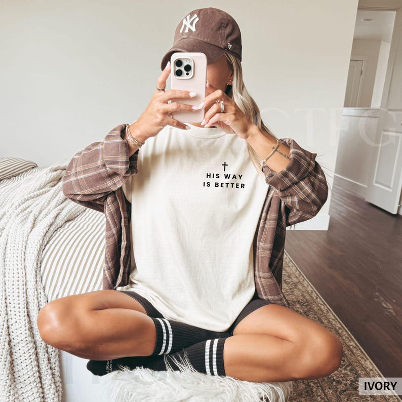 May include: An ivory-colored t-shirt with the text "HIS WAY IS BETTER" and a cross graphic. The person is wearing a brown baseball cap and a brown plaid shirt. The person is also wearing black socks with white stripes.
