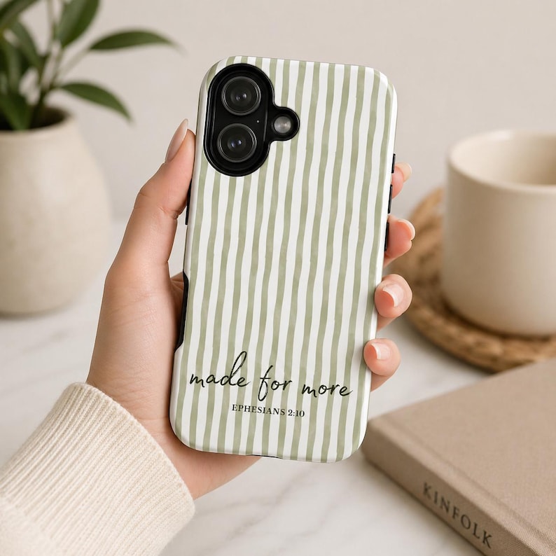 May include: A white and green striped phone case with the text "made for more" and "Ephesians 2:10" in black script. The case has a black border and is held in a person's hand. A white mug and a book are in the background.