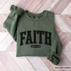 Puede incluir: Una sudadera verde militar con la palabra "FAITH" en letras negras grandes en la parte delantera. Debajo, el texto "MATT. 17:20". La sudadera est&aacute; doblada sobre una superficie de madera.