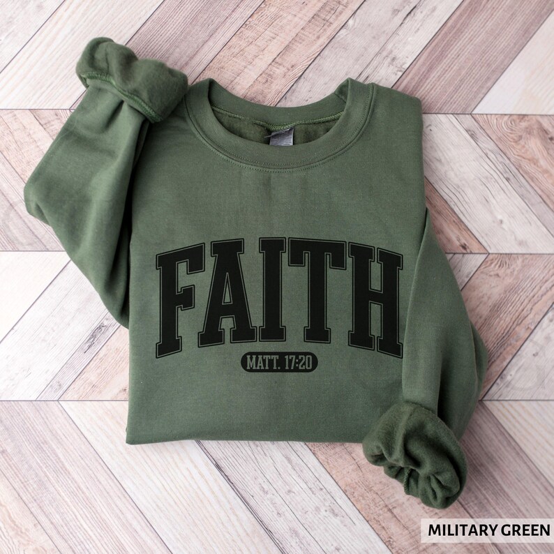 Puede incluir: Una sudadera verde militar con la palabra "FAITH" en letras negras grandes en la parte delantera. Debajo, el texto "MATT. 17:20". La sudadera est&aacute; doblada sobre una superficie de madera.