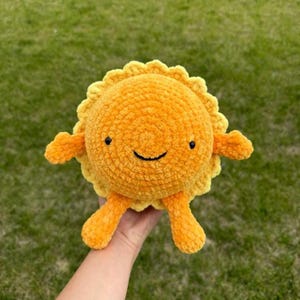 以下が含まれることがあります： 笑顔の顔をした、手作りのぬいぐるみのおもちゃ。太陽は主にオレンジ色で、黄色のスカラップエッジが付いています。小さな黒い目とオレンジ色の腕と脚があります。おもちゃは緑の草の背景に対して保持されています。