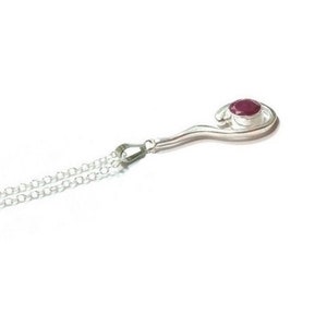 May include: A silver necklace featuring a delicate chain and a pendant with a smooth, curved metal frame. The pendant is set with a single, round, deep red gemstone at its center, creating a contrast between the polished silver and the stone.