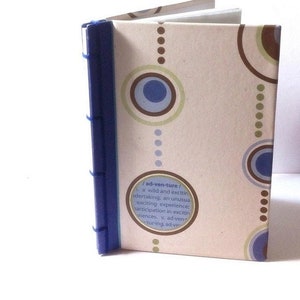 May include: A small, open notebook with a blue spine and a patterned cover. The cover features a beige background with blue, brown, and green circles and the word "adventure" with a definition.