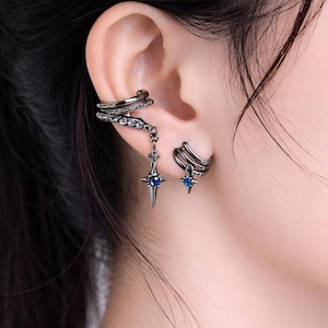 Könnte beinhalten: Silberfarbene Ohrklemmen und Ohrringe. Eine Ohrklemme umschließt das Ohr mit einem hängenden Sternanhänger mit einem blauen Edelstein. Eine weitere Ohrklemme hat ein schlichtes Design. Die Ohrringe haben ein Sterndesign mit einem blauen Edelstein.