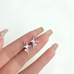 May include: A pair of silver star-shaped earrings. Each earring has a four-pointed star design with a smaller cross-like shape and a cluster of small, clear crystals. The earrings are shown against a light background.