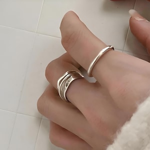Op de afbeelding: Twee zilveren ringen worden op een hand getoond. Eén is een eenvoudige, dunne band. De andere ring heeft een bredere band met een gelaagd, getextureerd ontwerp. De ringen zijn glanzend en reflecteren licht.