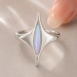 Puede incluir: Un anillo plateado con un diseño único. El anillo presenta un elemento central alargado en forma de diamante con una incrustación nacarada e iridiscente en tonos azules y morados. La banda del anillo es lisa y pulida.