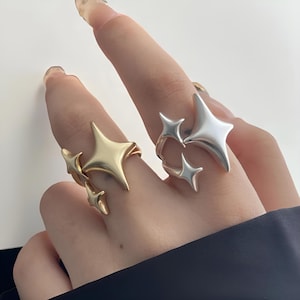 Puede incluir: Dos anillos, uno dorado y otro plateado, cada uno con un diseño de estrella. El anillo dorado tiene tres estrellas, mientras que el anillo plateado tiene tres estrellas. Los anillos se llevan en los dedos.