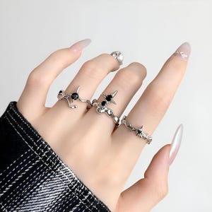 May include: A hand adorned with multiple silver rings, each featuring black gemstones. The rings showcase diverse designs, such as starburst and branch motifs. The rings are worn across several fingers, highlighting their intricate details and contrasting colours.