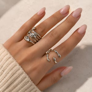 Könnte beinhalten: Zwei silberfarbene Ringe mit einer unregelmäßigen, geschmolzenen Metalltextur. Ein Ring hat ein breites, mehrschichtiges Design, während der andere ein dünnerer, offener Ring mit abgerundeten, fließenden Formen ist, die den Finger umschließen.