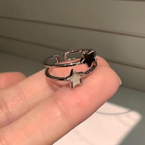 Op de afbeelding: Een zilverkleurige ring met twee sterontwerpen. Eén ster is zwart, de andere wit. De verstelbare ring zit op een vinger.