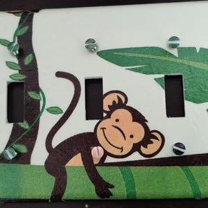 May include: A white light switch cover with a jungle theme. A brown monkey with a yellow face is climbing a vine. The background features green leaves and a brown tree trunk.