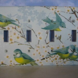May include: A light blue and yellow bird on a white background with a floral design. The bird is perched on a branch with white flowers. The background is a light blue with a white floral design. The image is a decorative light switch cover.