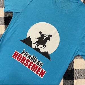 May include: A light blue t-shirt featuring a graphic of the Headless Horseman riding a horse in silhouette against a white moon. The words "Headless Horsemen" are printed in red and white.