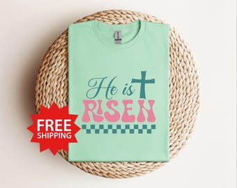 Könnte beinhalten: Ein hellgrünes T-Shirt mit dem Text "He is RISEN" in einer Retro-Schrift. Der Text ist in Türkis und Rosa mit einem türkisfarbenen Kreuz über dem Text. Unter dem Text befindet sich ein kariertes Muster.