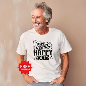 May include: Cream-colored t-shirt with the text "Botanical Beauty HAPPY DUTY" in black script. The design includes cactus illustrations and star accents. The shirt is worn by a person.