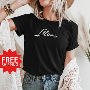 Puede incluir: Camiseta negra con texto blanco que dice "Illinois" en letra cursiva. La camiseta la lleva puesta una persona con el pelo largo y rubio.